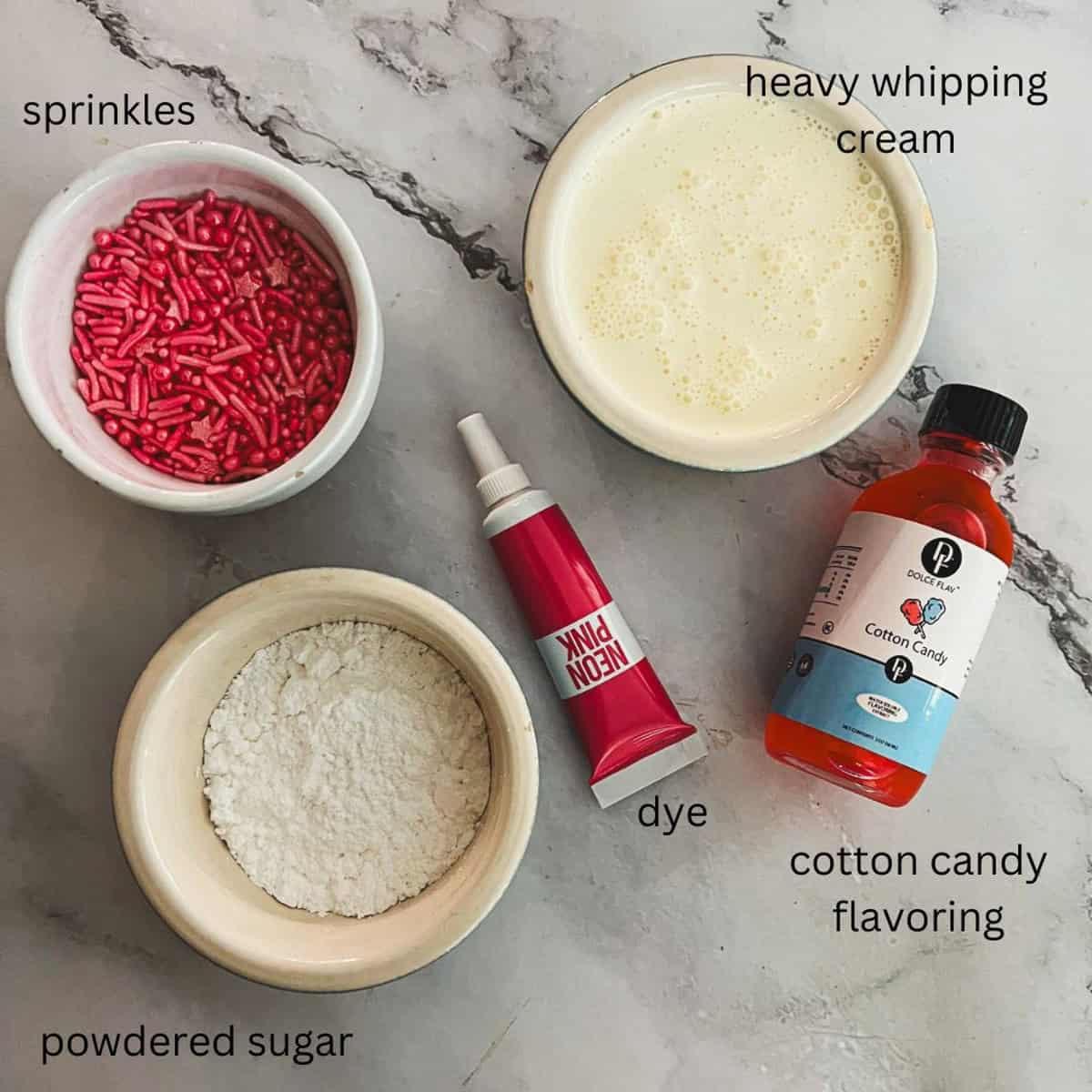 Four bowls on a marble surface containing the ingredients for cotton candy ingredients cream: a bowl of pink sprinkles, a bowl of heavy whipping cream, a bowl of powdered sugar, a tube of pink food dye, and a bottle of cotton candy flavoring.