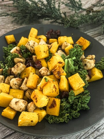 A plate of roasted butternut squash cubes, kale, chicken, and dried cranberries. The dish is garnished with a sprig of rosemary.