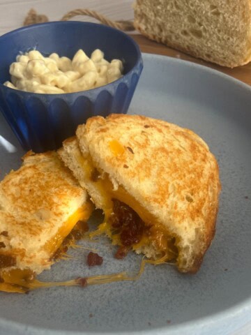 A delicious-looking sourdough grilled cheese sandwich with bacon bits and a side of macaroni and cheese.