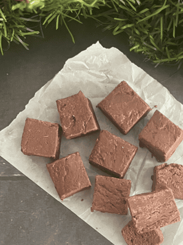 A batch of rich, dark chocolate fudge has been cut into squares and placed on a sheet of parchment paper. The fudge has a glossy, slightly textured surface