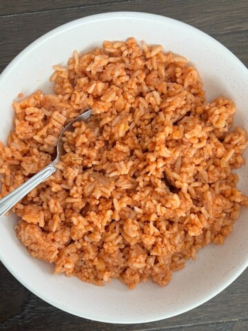 A white bowl filled with red rice sits on a dark wooden table. A silver spoon is resting in the Spanish rice box recipe.