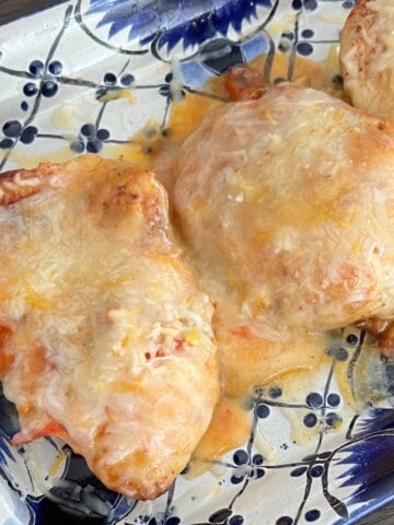 Three chicken breasts topped with melted cheese are arranged on a blue and white patterned platter as part of a Spanish chicken recipe.