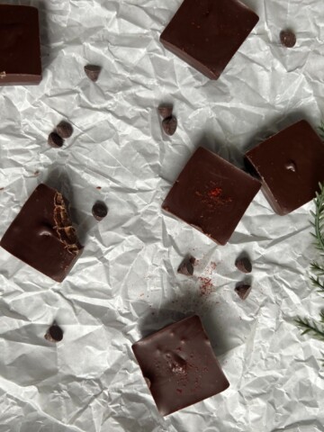 A top-down view of square pieces of dark spicy chocolate on a crumpled white surface. Some of the squares have a dusting of red chili powder and a few have chocolate chips scattered around them. A sprig of greenery and a few extra chocolate chips are visible in the bottom right corner.