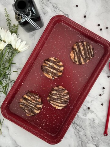 Four gluten-free strawberry coconut macaroons gluten free drizzled with chocolate on a red speckled enamel tray, with a red spoon and white daisies in the background.