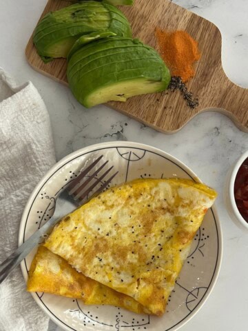 A plate of folded breakfast scrambled eggs seasoned with pepper, served with sliced avocado, a sprinkle of orange powder (possibly paprika), and a side of red salsa.