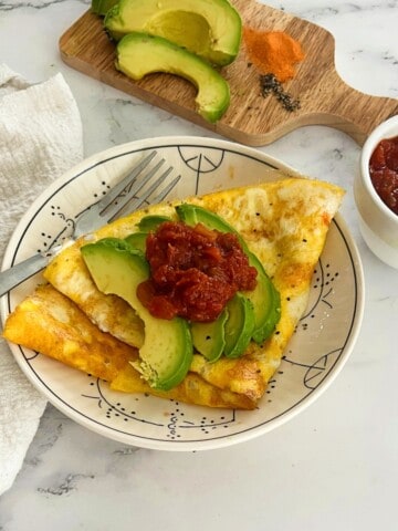 Top-down view of a white plate with a folded scrambled egg and avocado with salsa. The omelet is topped with slices of bright green avocado and a spoonful of chunky red salsa. A silver fork lies across the omelet. To the left of the plate is a folded white cloth napkin. In the upper right corner, a wooden cutting board holds half of a sliced avocado, a small pile of orange powder (possibly paprika), and a small pile of black peppercorns. A white bowl of red salsa is also on the cutting board. The scene is set on a light-colored marble countertop.