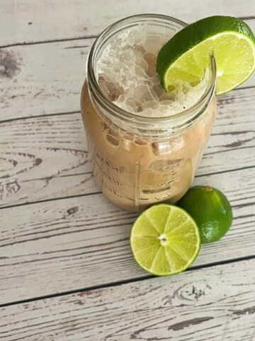 A mason jar filled with a Sonic-style Dirty Dr Pepper sits on a rustic white wooden surface. The drink is dark brown, filled with ice, and has visible carbonation bubbles on top. A fresh lime wedge is perched on the rim of the jar, while two additional limes, one whole and one sliced in half, rest nearby on the table. The background features a weathered wood texture, adding to the casual, homemade aesthetic.