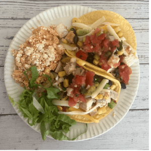 A vibrant image of two swordfish tacos on yellow corn tortillas, generously filled with grilled swordfish, black beans, corn salsa, shredded cheese, and fresh pico de gallo. They are served on a white paper plate, accompanied by a side of Mexican rice and a small bed of fresh green lettuce.