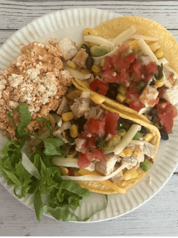 A vibrant image of two swordfish tacos on yellow corn tortillas, generously filled with grilled swordfish, black beans, corn salsa, shredded cheese, and fresh pico de gallo. They are served on a white paper plate, accompanied by a side of Mexican rice and a small bed of fresh green lettuce.