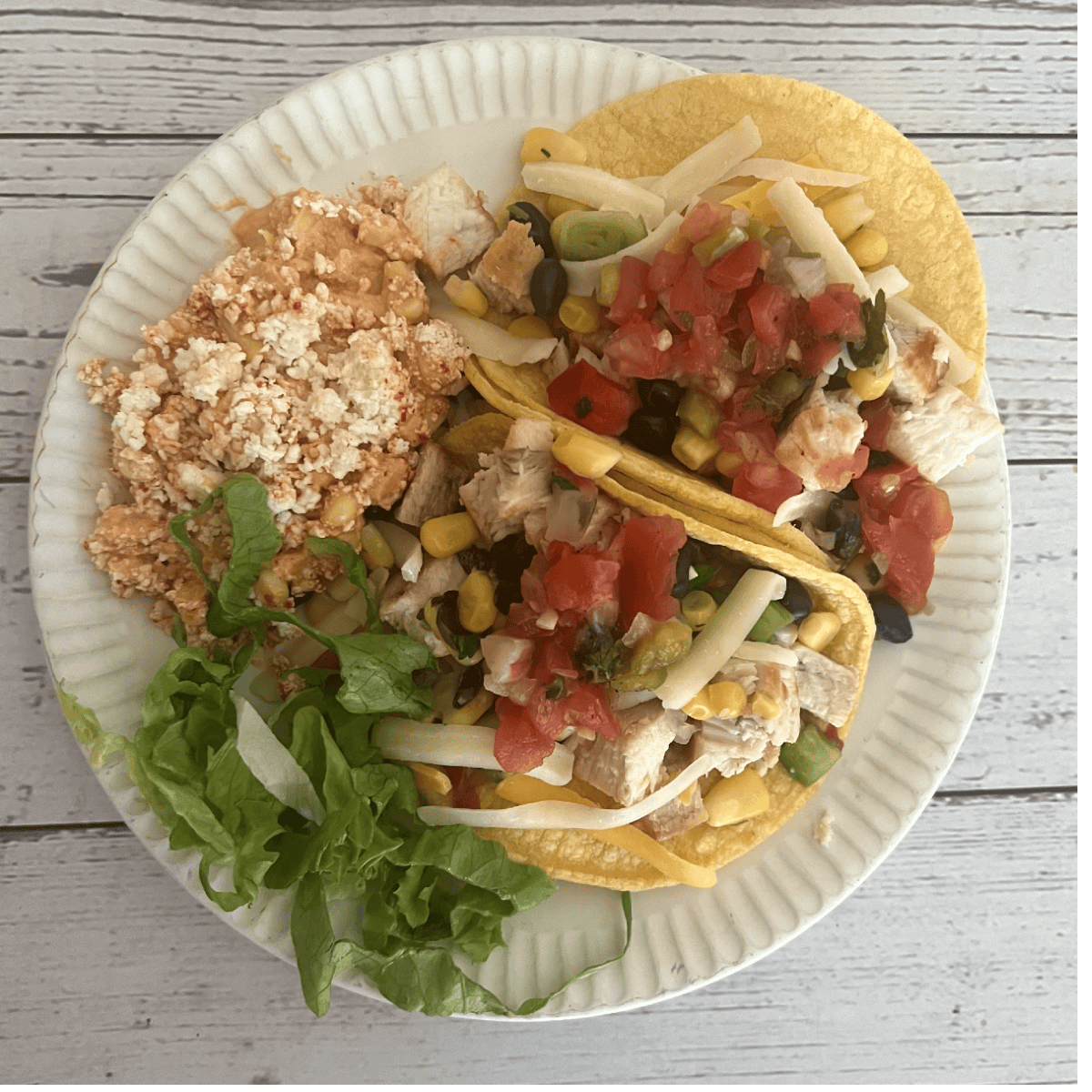 A vibrant image of two swordfish tacos on yellow corn tortillas, generously filled with grilled swordfish, black beans, corn salsa, shredded cheese, and fresh pico de gallo. They are served on a white paper plate, accompanied by a side of Mexican rice and a small bed of fresh green lettuce.