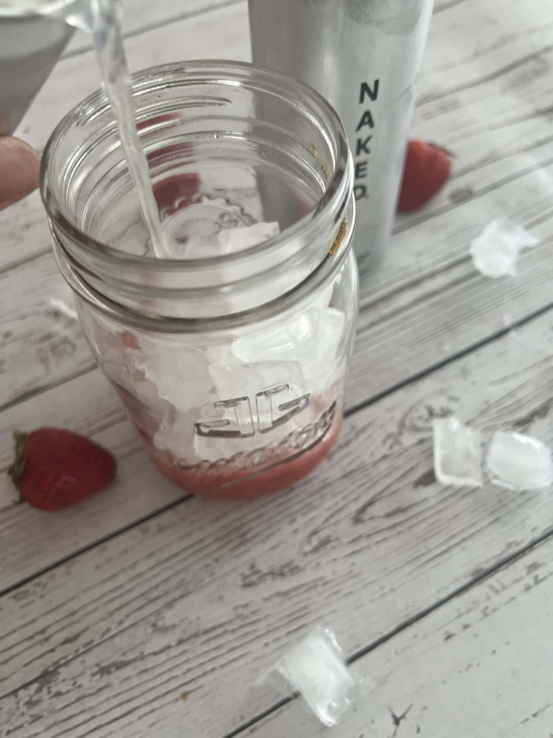 A liquid is being poured into a clear glass jar filled with ice cubes and a few strawberries on a weathered white wooden surface. In the background, a silver can of Naked brand Strawberry Lemonade sparkling energy drink and scattered ice cubes are visible.