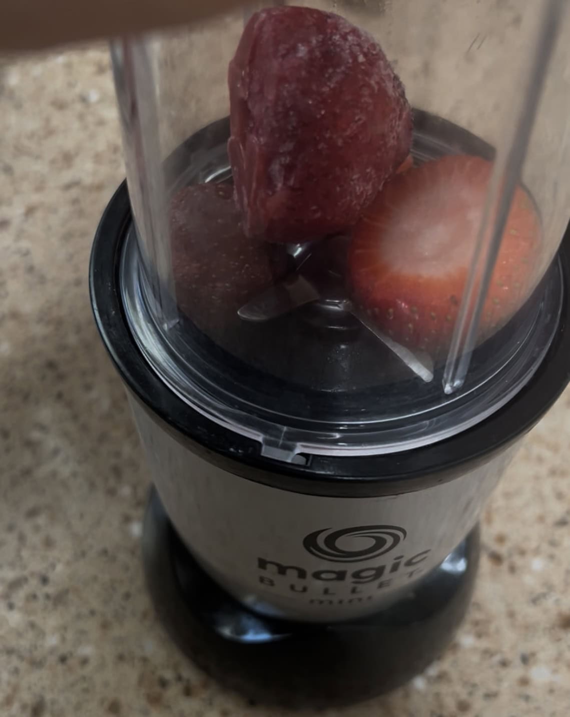 A small Magic Bullet blender cup with a black base contains three strawberries—one whole and two halved—resting on the blades. The transparent cup is ready to be blended.