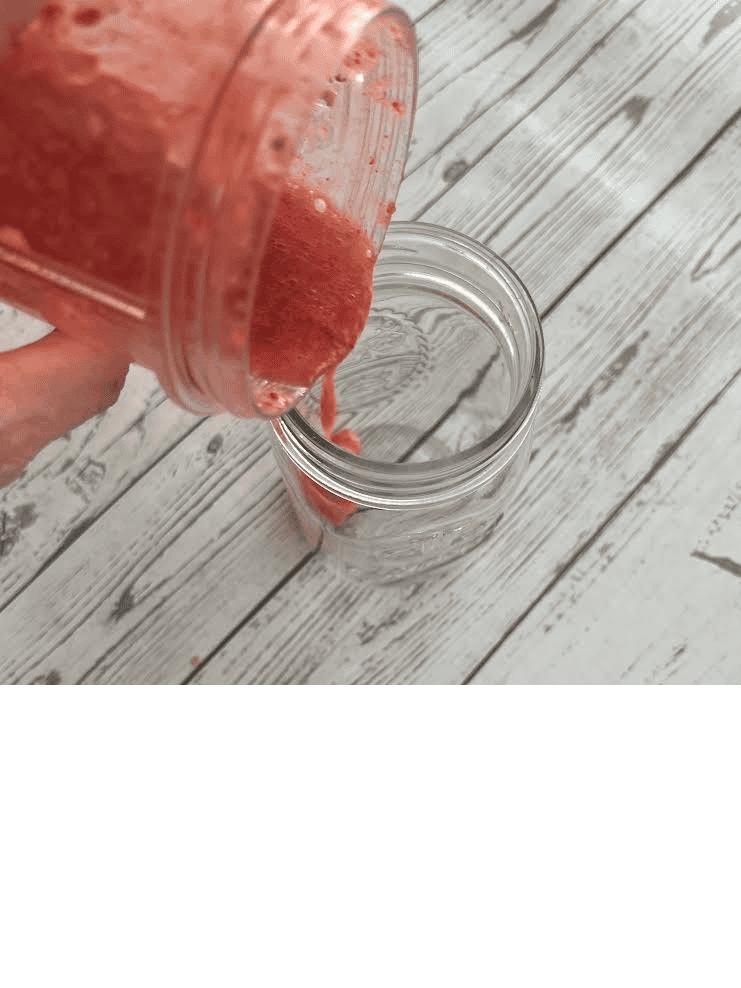 A thick, bright red strawberry purée is being poured from a Magic Bullet blender cup into a clear glass jar on a white weathered wooden table.