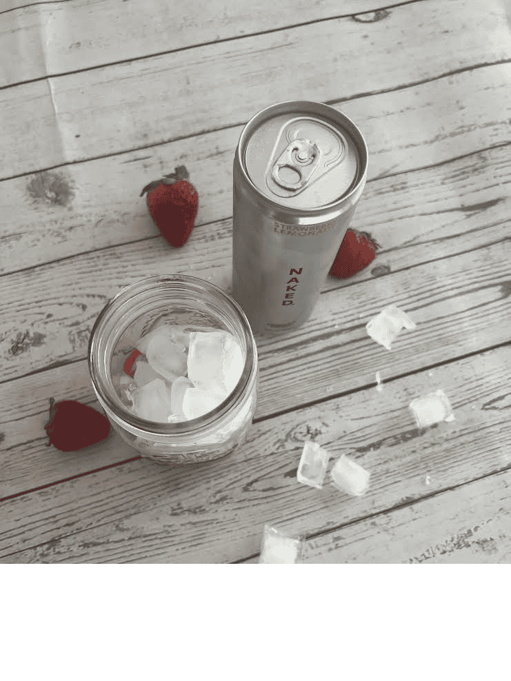 A glass jar with ice cubes and a few strawberries at the bottom sits on a weathered white wooden surface. Next to it is a closed can of Naked brand Strawberry Lemonade sparkling energy drink.