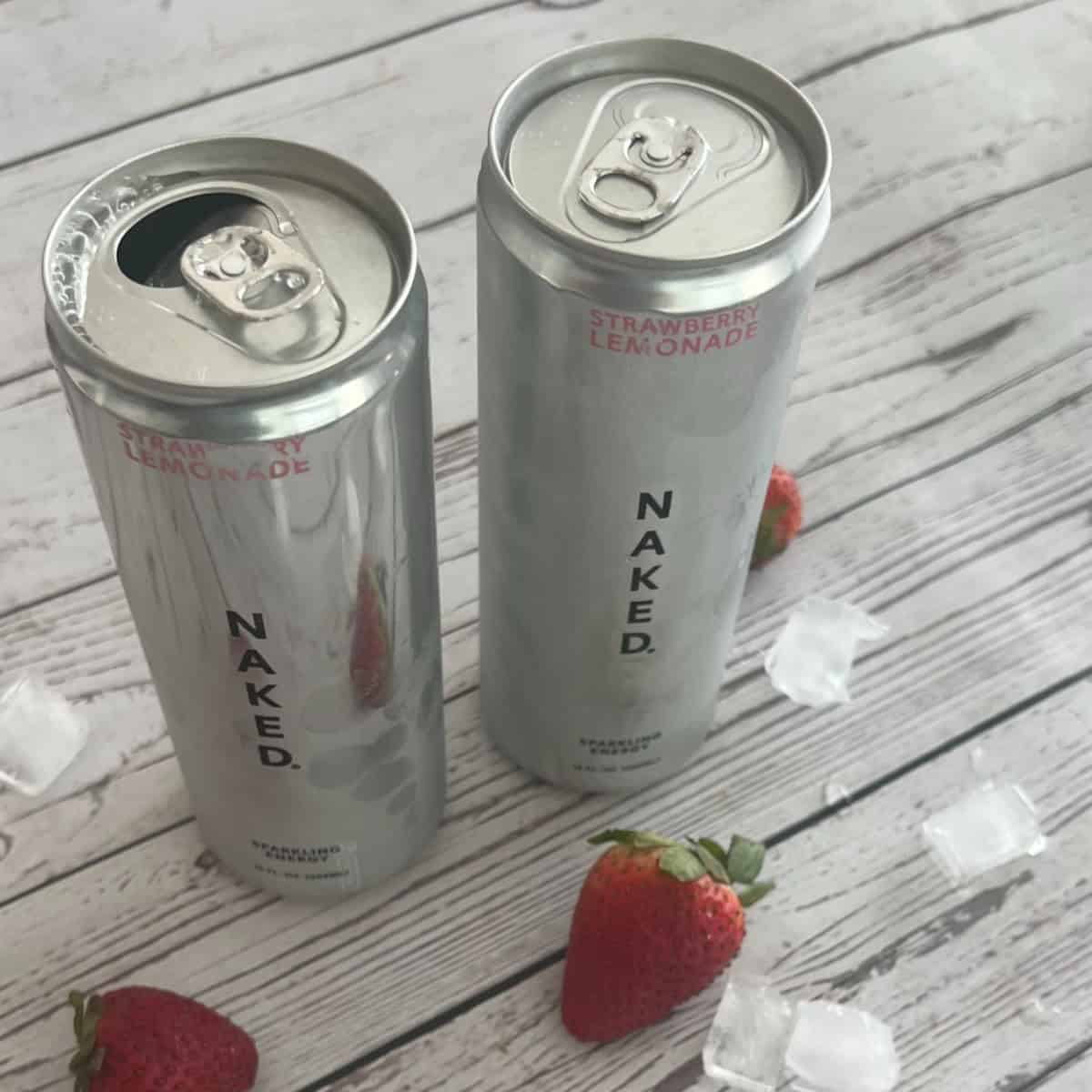 A glass jar with a pink strawberry lemonade drink and ice sits on a white wooden surface, next to a silver can of "NAKED." brand strawberry lemonade clean energy drink. Scattered around are ice cubes and a few fresh strawberries.
