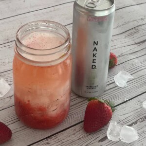 A chilled glass jar filled with a pink drink and ice sits on a weathered white wooden surface, alongside a silver can of "NAKED nutrition." brand strawberry lemonade sparkling energy drink. Scattered around the jar and can are individual ice cubes and a few fresh red strawberries.