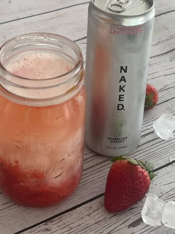 A chilled glass jar filled with a pink drink and ice sits on a weathered white wooden surface, alongside a silver can of "NAKED nutrition." brand strawberry lemonade sparkling energy drink. Scattered around the jar and can are individual ice cubes and a few fresh red strawberries.