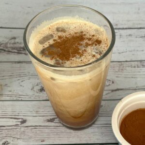 A clear glass of Starbucks Iced Brown Sugar Oatmilk Shaken Espresso sits on a weathered light wood surface. The drink, a mix of light brown and creamy white, is topped with foam and a dusting of dark cinnamon. A small white bowl filled with cinnamon powder is next to the glass. The recipe is for a skinny Starbucks shaken espresso recipe.
