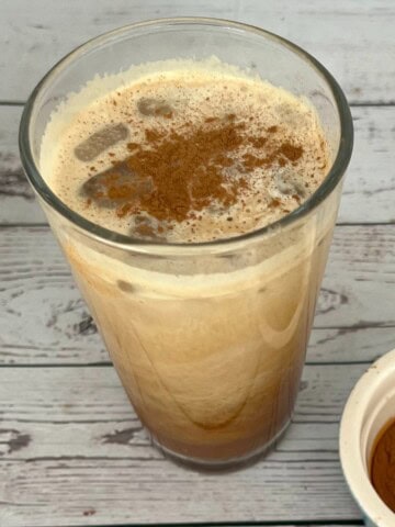 A clear glass of Starbucks Iced Brown Sugar Oatmilk Shaken Espresso sits on a weathered light wood surface. The drink, a mix of light brown and creamy white, is topped with foam and a dusting of dark cinnamon. A small white bowl filled with cinnamon powder is next to the glass. The recipe is for a skinny Starbucks shaken espresso recipe.