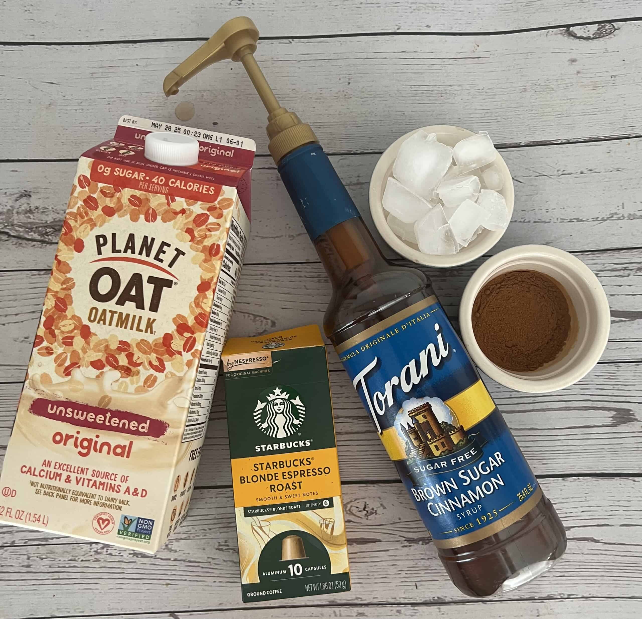 A high-angle shot shows the ingredients for a coffee drink laid out on a light wood surface. From left to right, there is a carton of Planet Oat Unsweetened Original Oatmilk, a box of Starbucks Blonde Espresso Roast Nespresso capsules, a bottle of Torani Sugar Free Brown Sugar Cinnamon Syrup, a small white bowl of ice cubes, and a small white bowl of ground cinnamon.