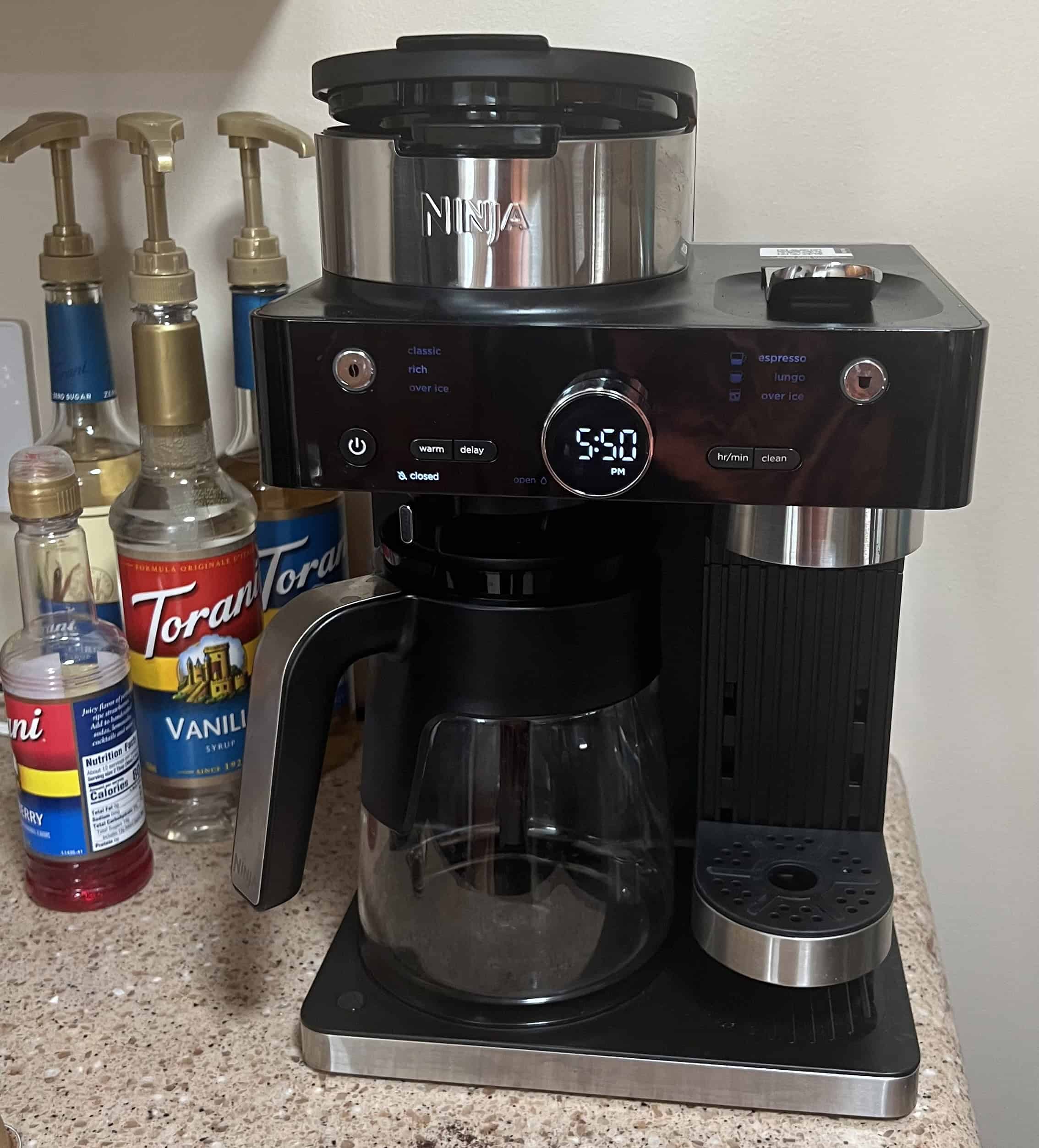 A full-length, eye-level shot of a Ninja coffee maker on a speckled countertop. The coffee maker is black with silver accents. It has a glass carafe and a built-in milk frother on the right. To the left of the coffee maker are several syrup bottles, including Torani Vanilla and a sugar-free vanilla syrup. The time on the coffee maker's display shows "5:50". The coffee maker appears clean and ready for use.