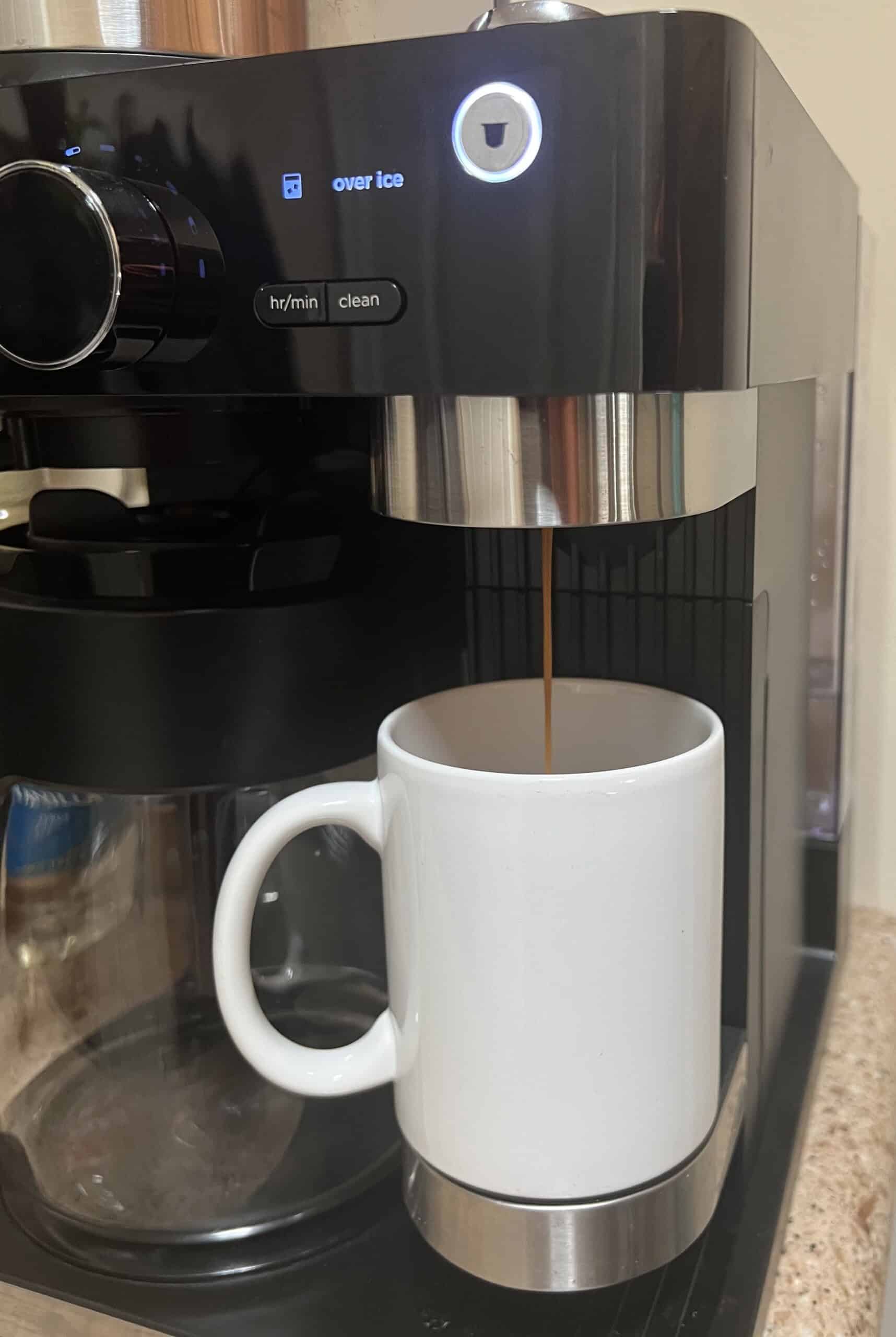 A vertical, eye-level close-up shot of a Ninja coffee maker. A thin stream of coffee is pouring from the machine's spout into a plain white mug. The coffee maker's digital screen displays the words "over ice" and an icon of a cup. The background is a speckled countertop.