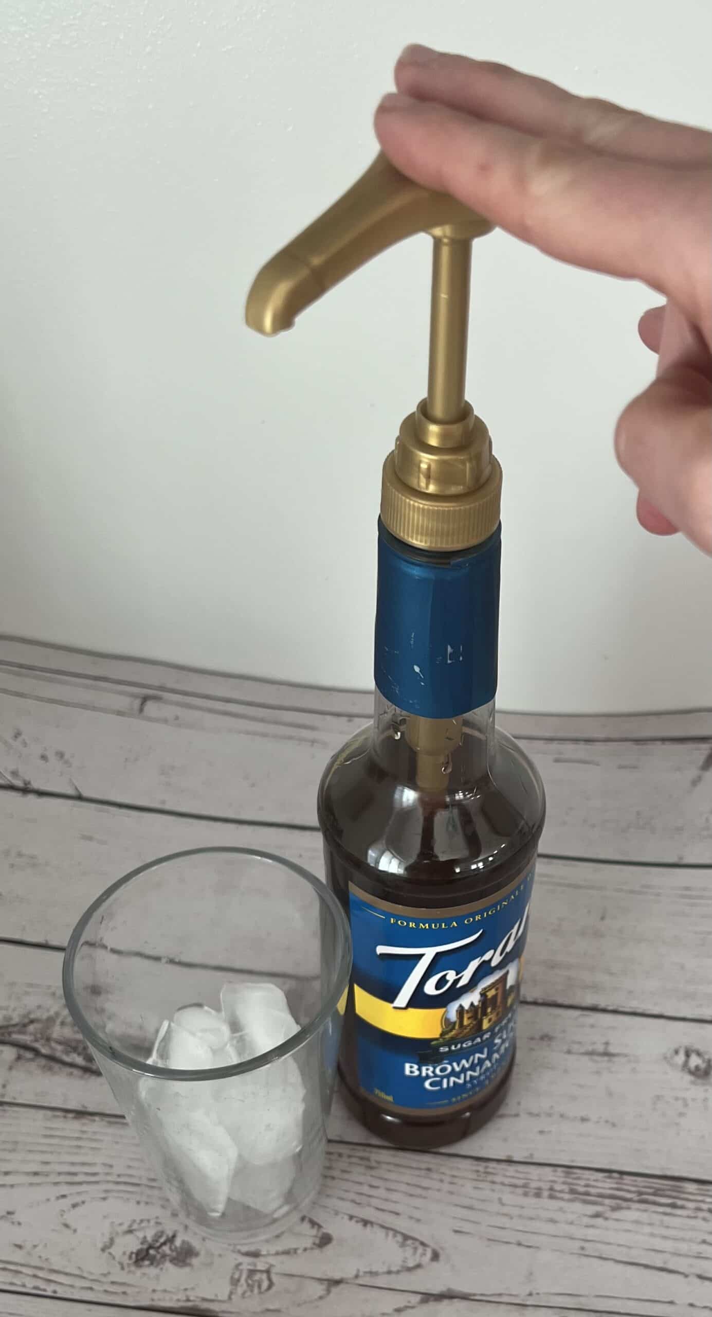 A hand is shown from the side, pressing down on a golden pump attached to a bottle of Torani Brown Sugar Cinnamon syrup. Next to the bottle is a clear glass with ice cubes inside. The items are on a light-colored wooden surface against a plain white wall.