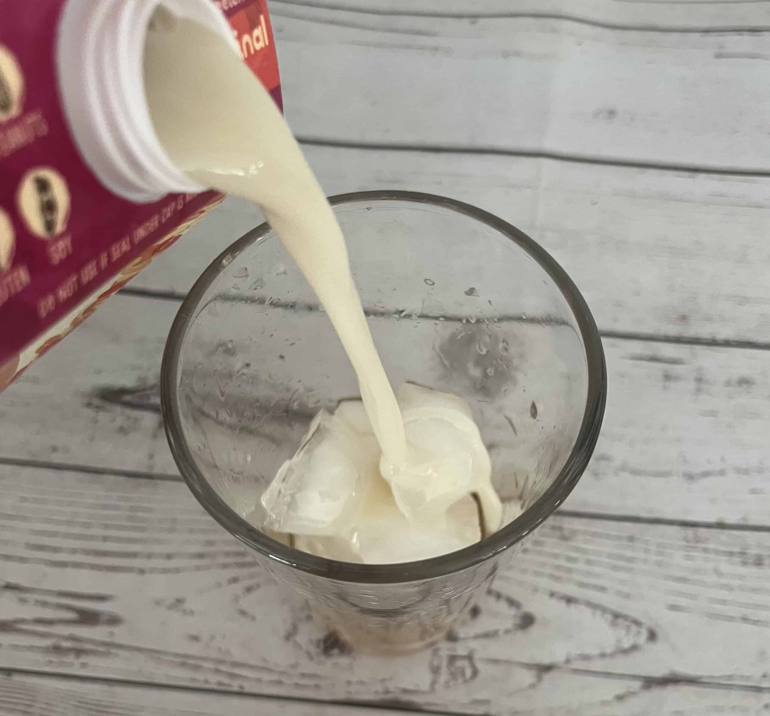 A vertical, close-up shot of creamy liquid oatmilk being poured from a pink and white container into a clear glass with ice. The liquid is hitting the ice, splashing slightly inside the cup. The container's label is partially visible on the left side, showing a red "original" sticker. The items are on a light wooden surface against a plain white wall.